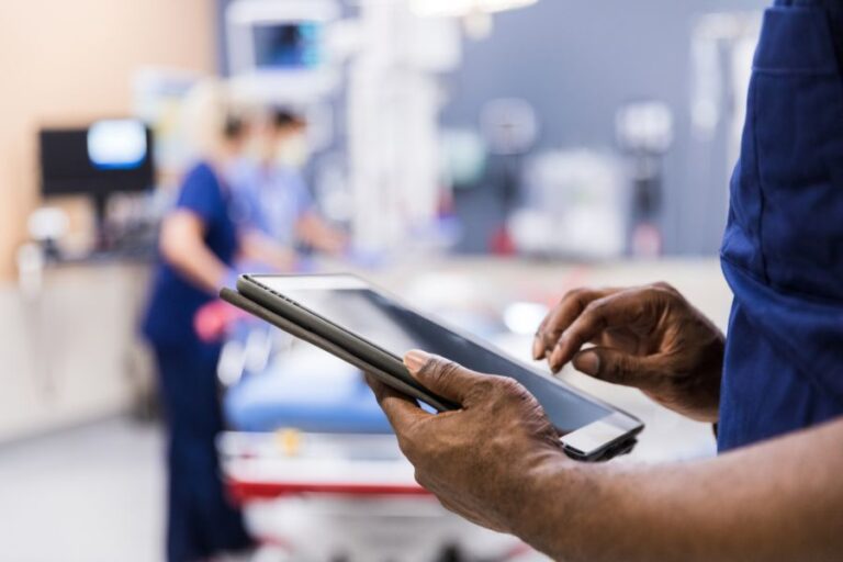 A healthcare professional uses a tablet in the foreground, while two other professionals are seen working in the background. The setting appears to be a medical facility.