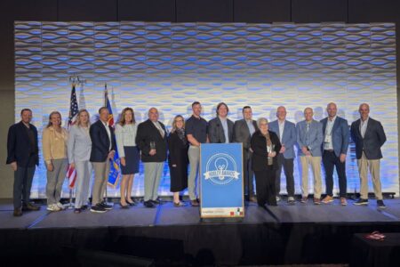 A group of sixteen people stands on a stage in front of flags and a sign reading "MOBILE AWARDS," posing for a group photo at an event.