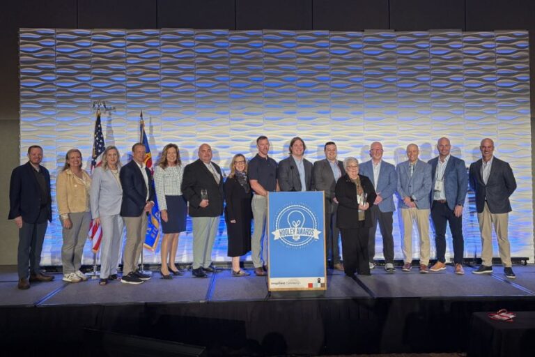 A group of sixteen people stands on a stage in front of flags and a sign reading "MOBILE AWARDS," posing for a group photo at an event.