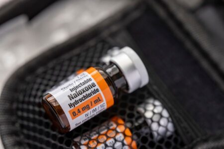 A vial labeled "Injectable Naloxone Hydrochloride 0.4 mg/mL" rests in a black zippered case with other medical supplies in the background.