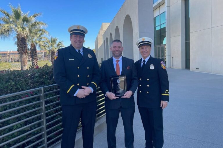 Three men, two in fire department uniforms and one in a suit holding a plaque, stand outside a modern building with palm trees in the background.