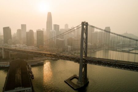 Aerial view of a bridge and city skyline under hazy skies, with the sun partially obscured and reflecting on the water below.