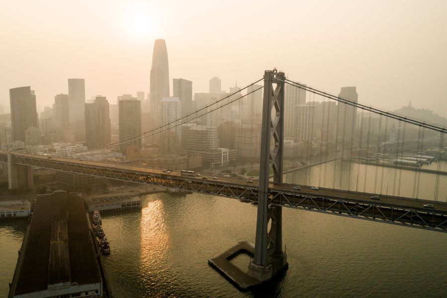 Aerial view of a bridge and city skyline under hazy skies, with the sun partially obscured and reflecting on the water below.