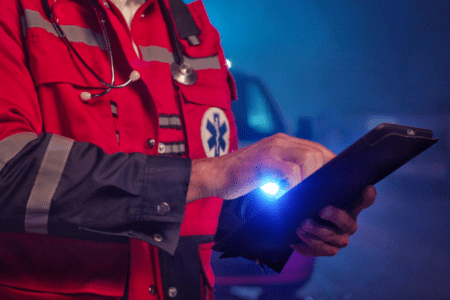 A paramedic in a red uniform uses a tablet during nighttime, with a stethoscope around their neck and an emergency vehicle in the background.