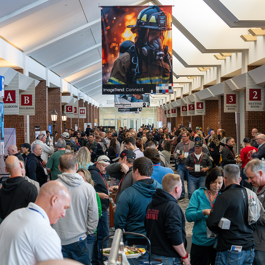 A large crowd gathers at ImageTrend Connect 25 in a convention center hallway beneath a banner featuring a firefighter in gear; numbered meeting rooms and ballrooms are visible in the background.