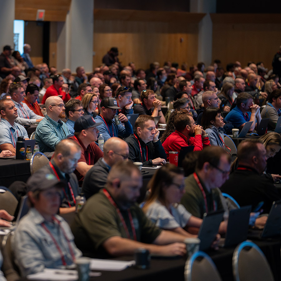 A large group of people sits in rows at a conference, listening attentively to a presentation. Many are using laptops or taking notes.