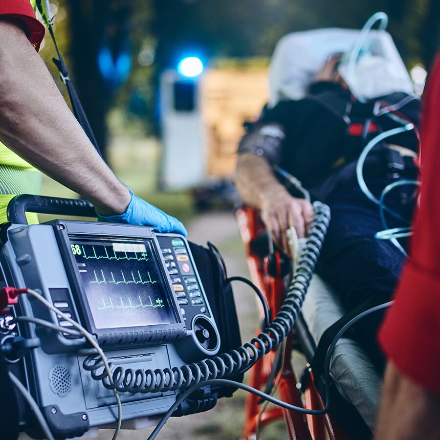 Paramedic prepares a defibrillator while a patient lies on a stretcher with medical monitoring equipment, outdoors near an ambulance.