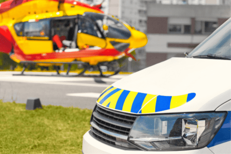 A police vehicle is parked in the foreground with a yellow and red rescue helicopter blurred in the background on a helipad.