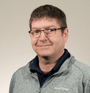 A man with short brown hair and glasses wearing a gray zip-up jacket over a dark shirt against a plain light background.