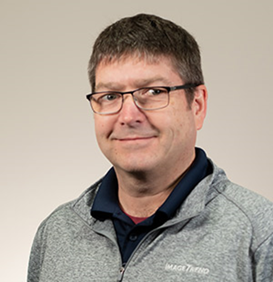 A man with short brown hair and glasses wearing a gray zip-up jacket over a dark shirt against a plain light background.