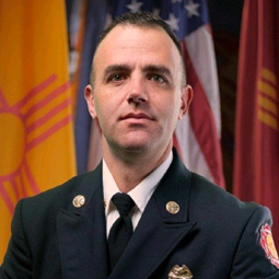 A uniformed firefighter poses for a formal portrait in front of U.S. and state flags.