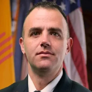 A man in a suit poses for a formal portrait with U.S. and state flags in the background.