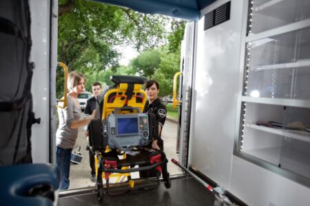 Two paramedics and a woman load a stretcher with medical equipment into an ambulance, viewed from inside the vehicle.