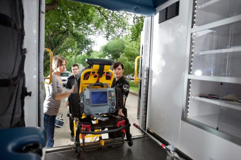 Two paramedics and a woman load a stretcher with medical equipment into an ambulance, viewed from inside the vehicle.