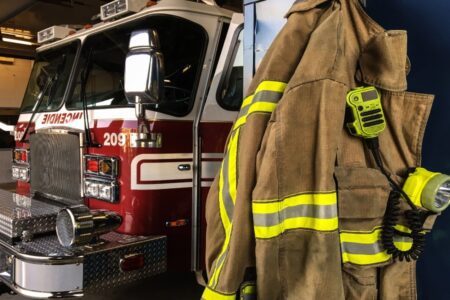 A fire truck is parked in a garage next to a hanging firefighter's turnout jacket equipped with a radio and flashlight.