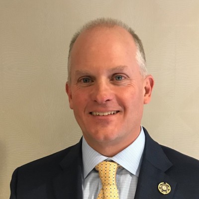 A man wearing a navy suit, light blue shirt, and yellow patterned tie stands in front of a beige background, smiling at the camera.
