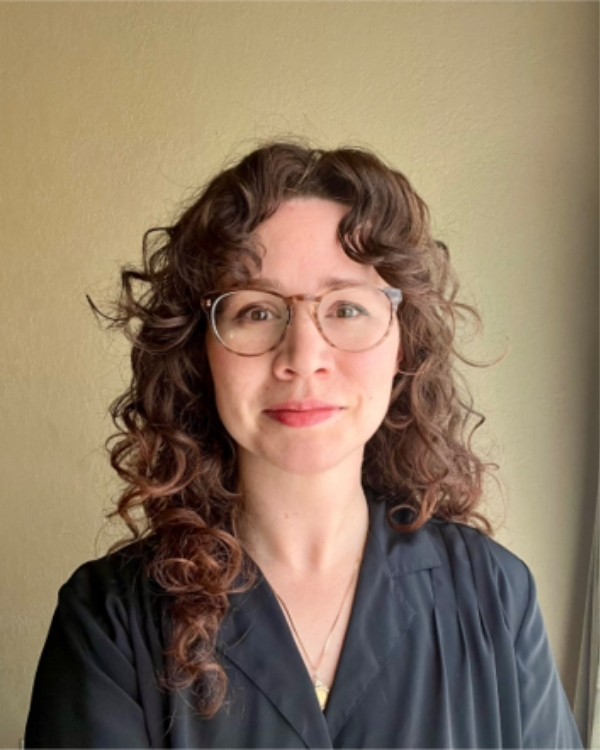 A woman with curly brown hair and glasses is standing indoors in front of a beige wall, wearing a dark top and smiling softly at the camera.