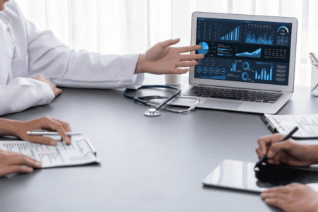 Doctors sitting at a table discuss medical data displayed on a laptop screen, with charts and graphs visible; tablets and documents are on the table.