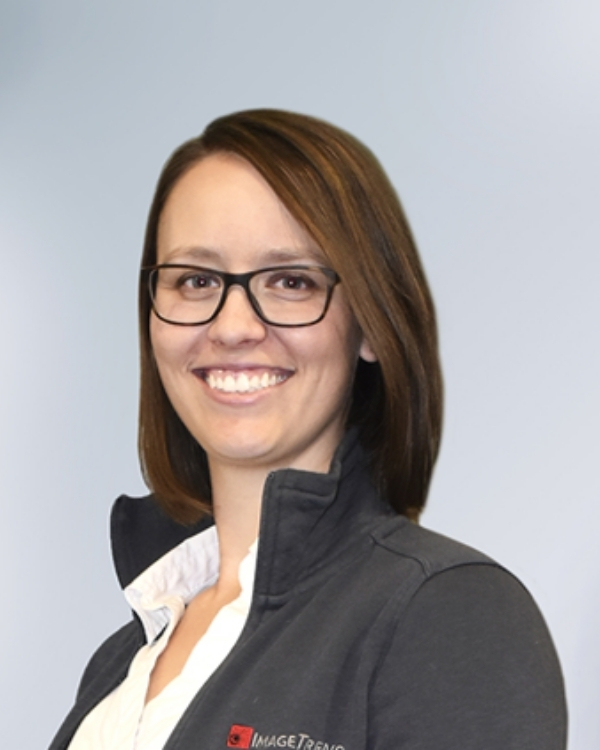 A woman with straight brown hair, wearing glasses and a dark zip-up jacket over a white shirt, smiles at the camera against a plain, light background.