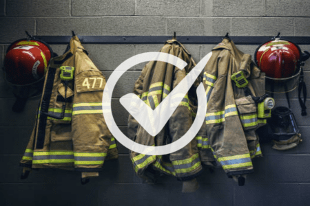 Three firefighter uniforms and helmets hang on hooks against a gray brick wall, with a large white check mark overlayed in the center.