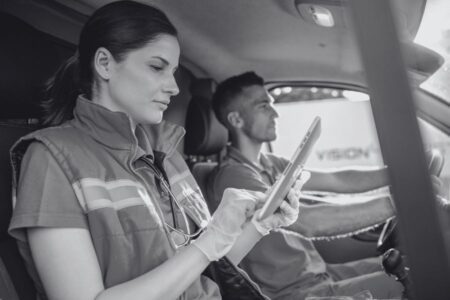 A woman wearing medical gloves uses a tablet in the passenger seat of a vehicle while a man drives.