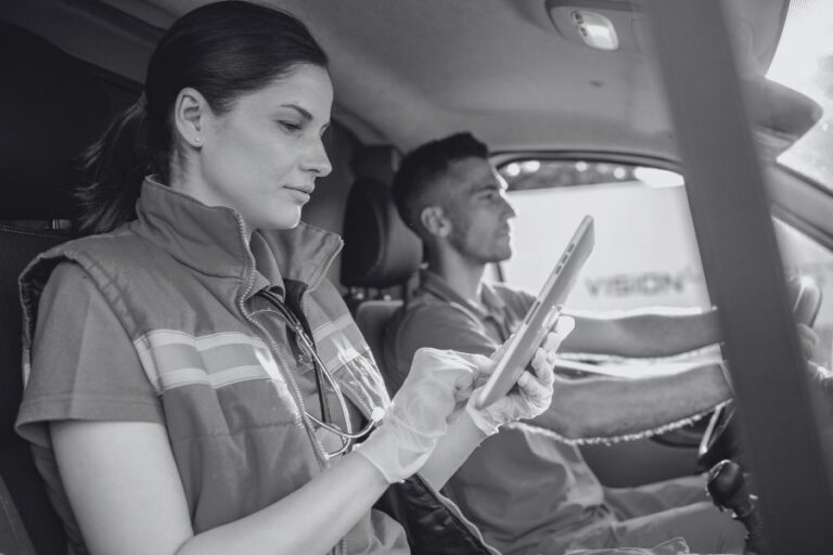 A woman wearing medical gloves uses a tablet in the passenger seat of a vehicle while a man drives.