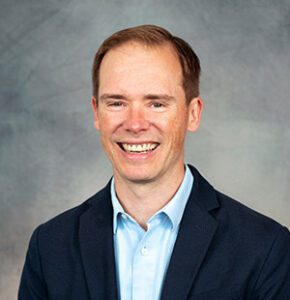 A man with short light brown hair is smiling, wearing a light blue shirt and dark blazer, posed in front of a gray background.