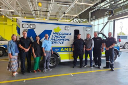 Eight people stand in front of a Middlesex London Paramedic Service ambulance inside a garage.