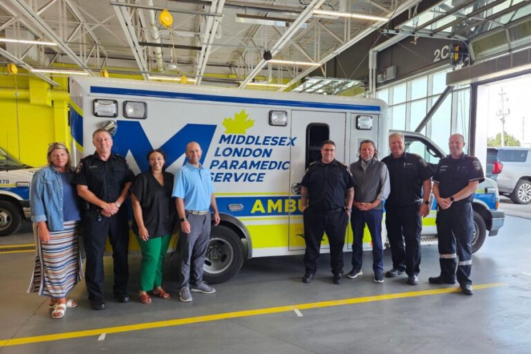 Eight people stand in front of a Middlesex London Paramedic Service ambulance inside a garage.