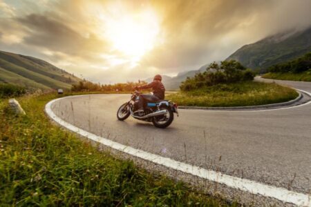 A person rides a motorcycle on a winding mountain road at sunset, surrounded by green hills and cloudy skies.