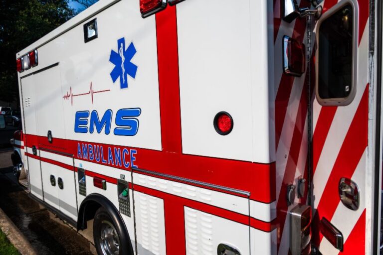 A white EMS ambulance with red stripes and medical symbols is parked on the side of a road.