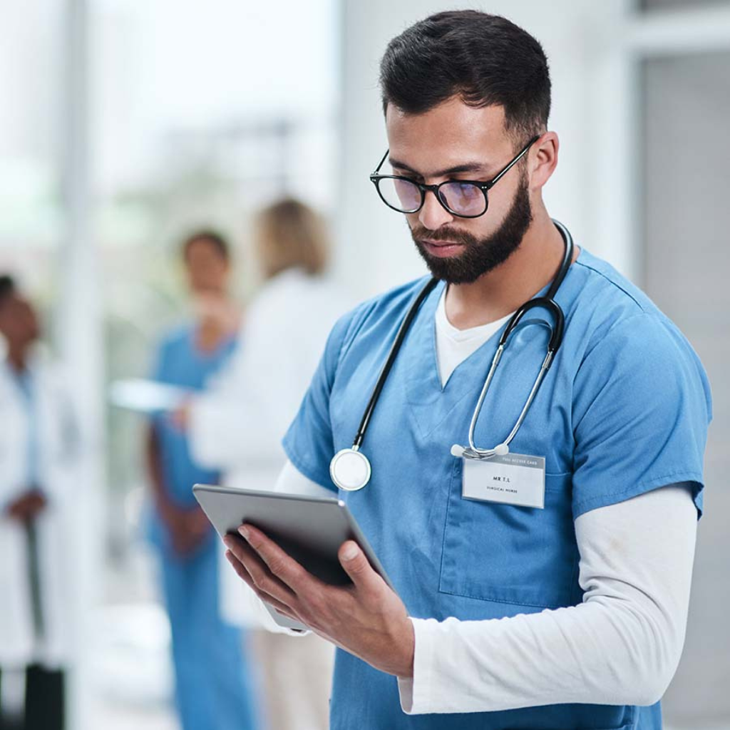 A male healthcare professional in blue scrubs and a stethoscope uses a tablet in a hospital setting, with other medical staff in the background.