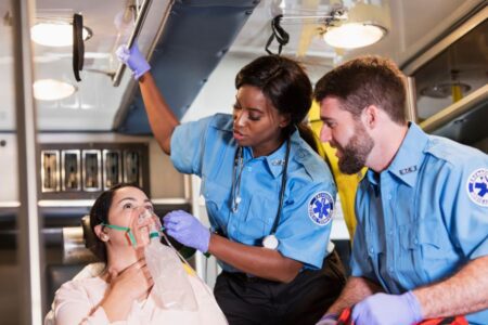 Two paramedics in uniform assist a patient with an oxygen mask in the back of an ambulance.