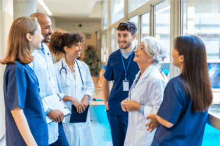 A group of healthcare professionals, including doctors and nurses, stand together in a hospital corridor, talking and smiling.