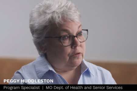 A woman with short gray hair and glasses, wearing a light blue shirt, is speaking. On-screen text identifies her as Peggy Huddleston, Program Specialist, MO Dept. of Health and Senior Services.