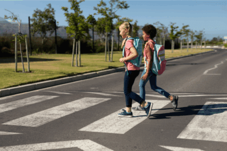 Two children with backpacks walk across a crosswalk on a sunny day, with trees and grass visible along the side of the road.