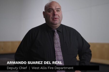 A man in a black shirt and patterned tie stands indoors. Text overlay reads: "Armando Suarez Del Real, Deputy Chief, West Allis Fire Department.