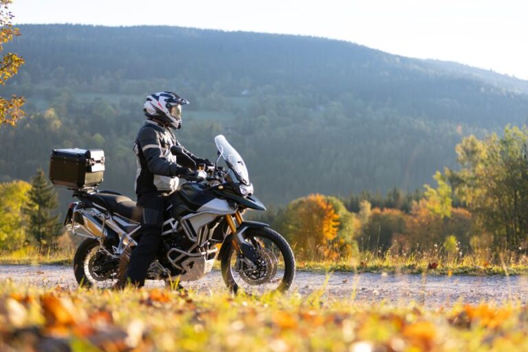 A person wearing motorcycle gear sits on an adventure touring motorcycle on a gravel path, with a scenic forested hillside in the background.