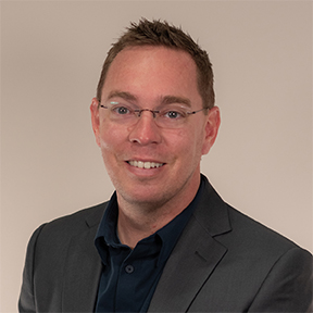 A man wearing glasses, a dark blazer, and a navy shirt smiles at the camera against a plain light background.