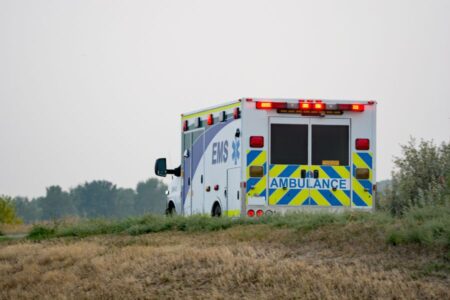 An ambulance with flashing lights drives on a rural road bordered by grass and bushes, with trees visible in the background.