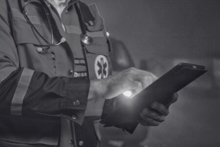 A person in a paramedic uniform uses a tablet device, with a stethoscope around their neck and an ambulance visible in the background.