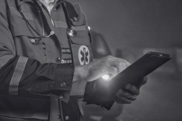 A person in a paramedic uniform uses a tablet device, with a stethoscope around their neck and an ambulance visible in the background.
