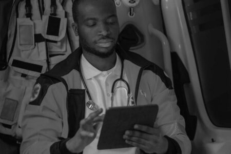A healthcare professional wearing a stethoscope sits inside an ambulance and uses a tablet device.
