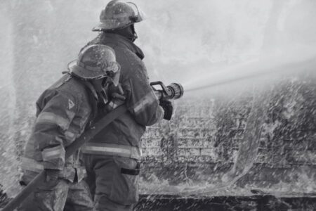 Two firefighters in protective gear spray water from a hose onto a fire, with water and mist surrounding them.