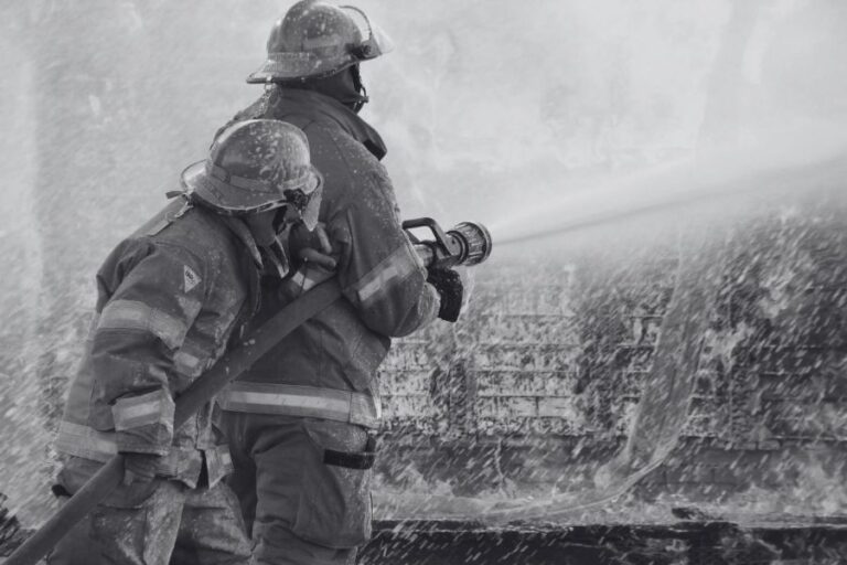 Two firefighters in protective gear spray water from a hose onto a fire, with water and mist surrounding them.