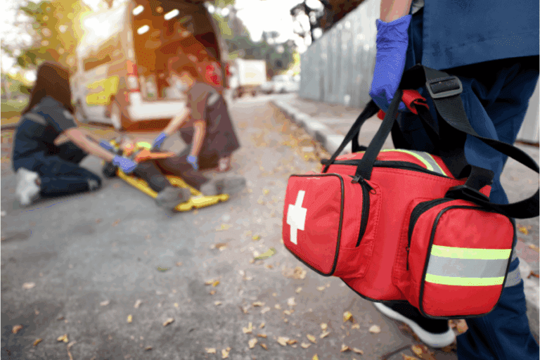 Paramedics attend to a person lying on the ground next to an ambulance, while another arrives carrying a red medical bag.