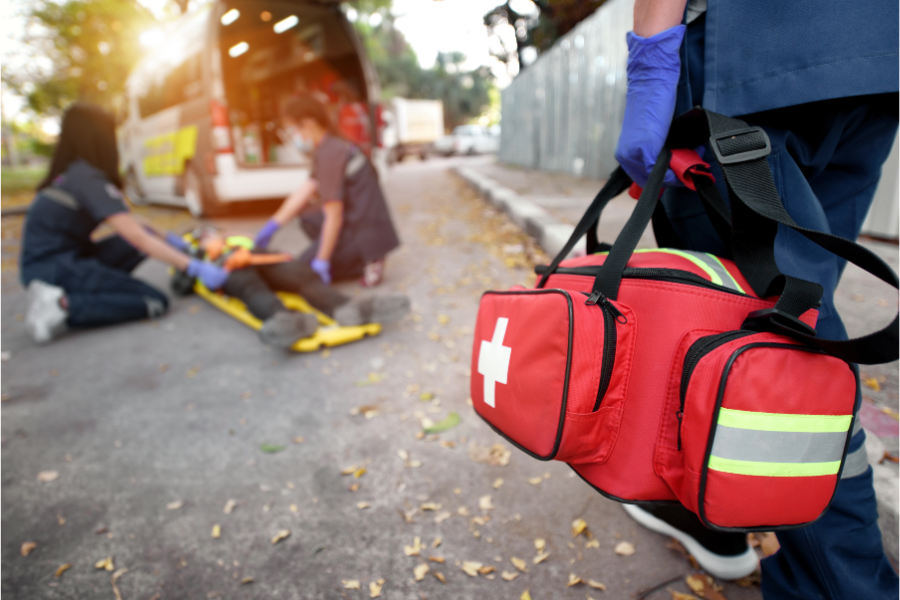 Paramedics attend to a person lying on the ground next to an ambulance, while another arrives carrying a red medical bag.