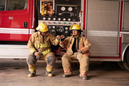Two firefighters in gear sit next to a fire truck, holding water bottles and fist-bumping in front of the vehicle’s control panel.
