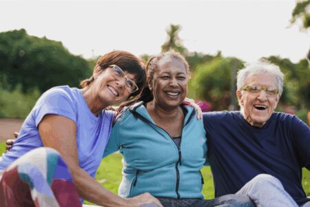 Three older adults sitting outdoors on grass, smiling and posing together with arms around each other. Trees and greenery are visible in the background.