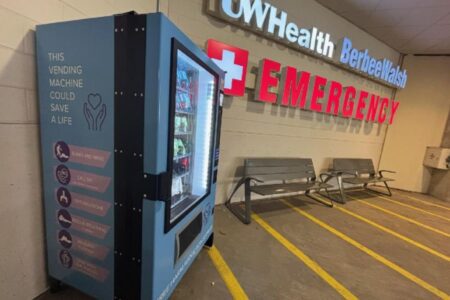 A vending machine with harm reduction supplies is placed near the entrance to an emergency room at UW Health, next to empty benches and emergency signs.